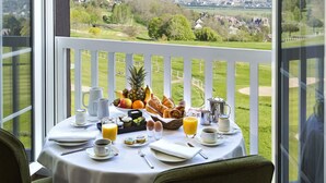 Una mesa de desayuno con diversos alimentos y bebidas, ubicada en un balcón con vista a un campo de golf y un paisaje urbano.