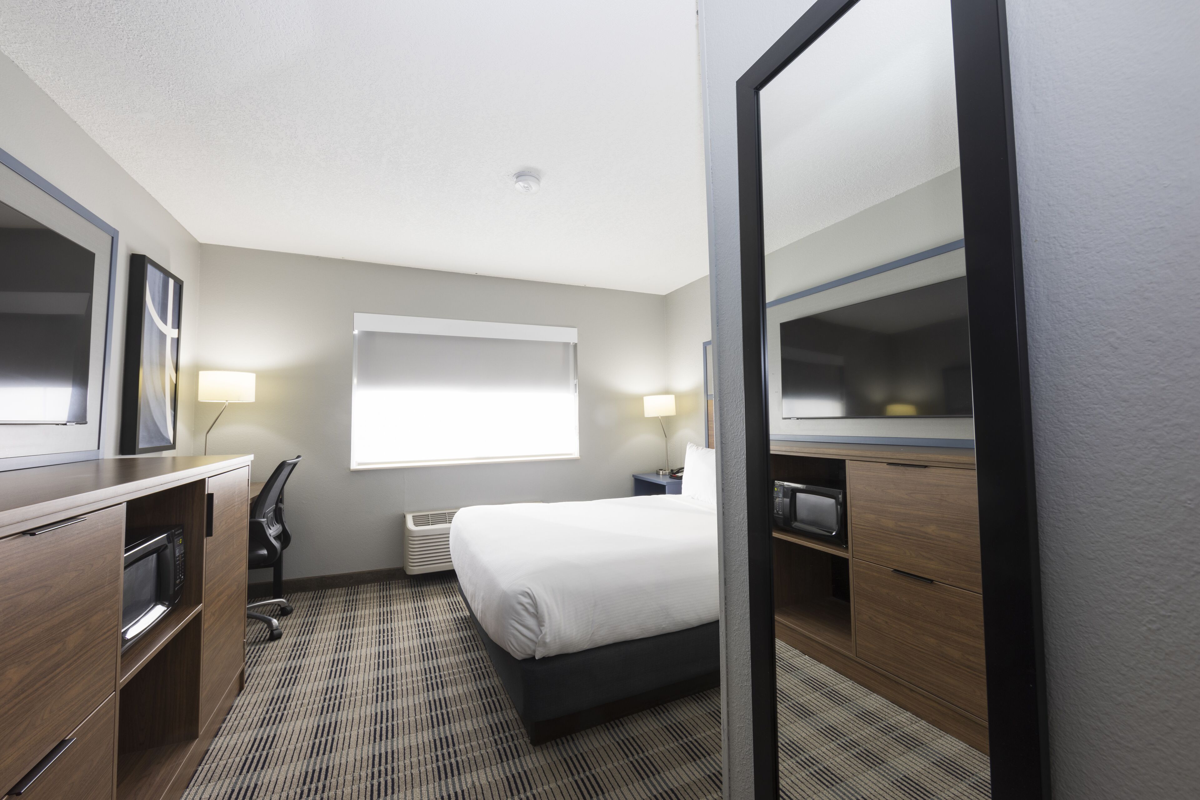 Habitación de hotel con cama, escritorio con microondas, silla, mesita de noche y un espejo grande.