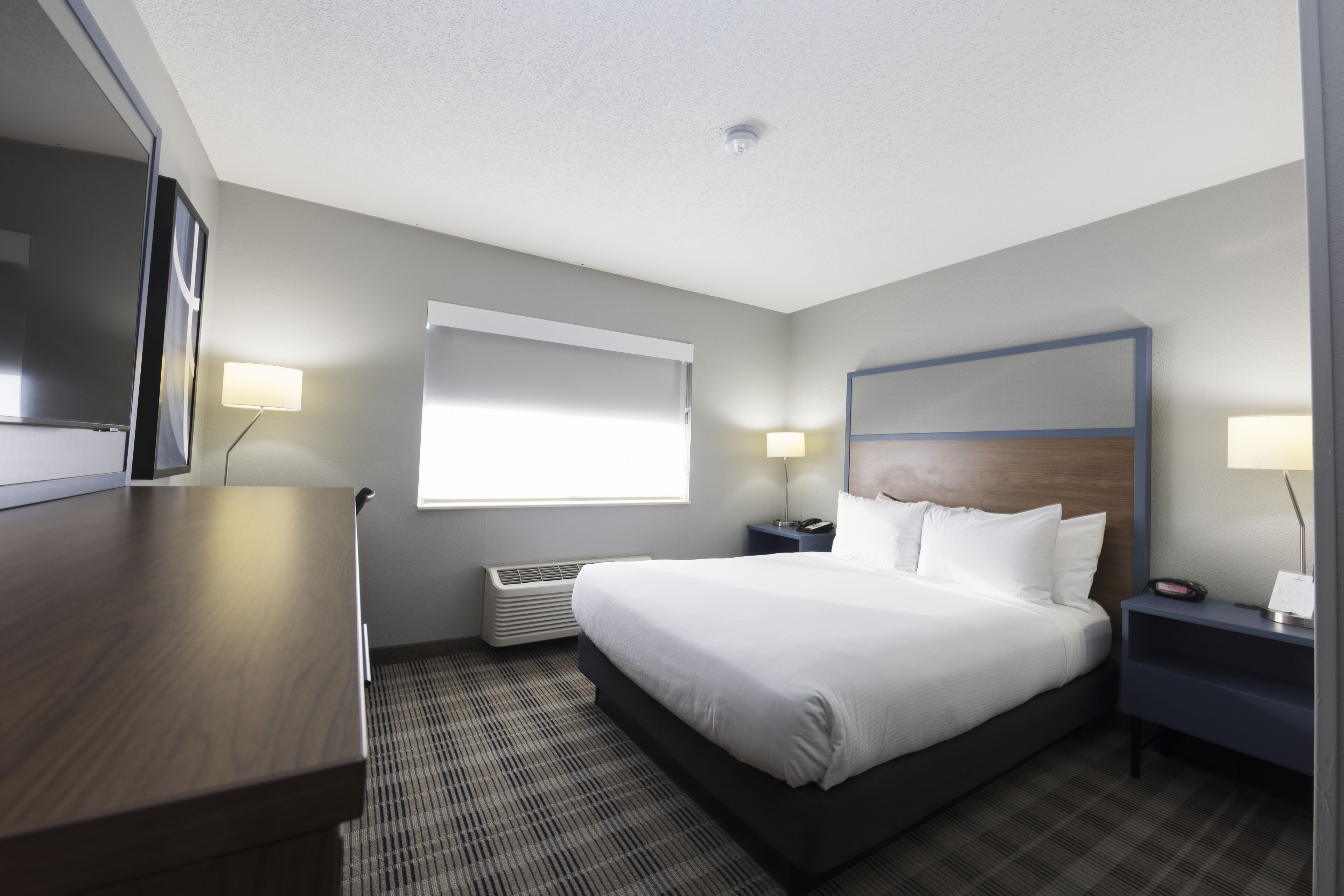 A hotel room with a large bed, a desk, two bedside tables, a flat-screen TV, a window with blinds, and a ceiling light.