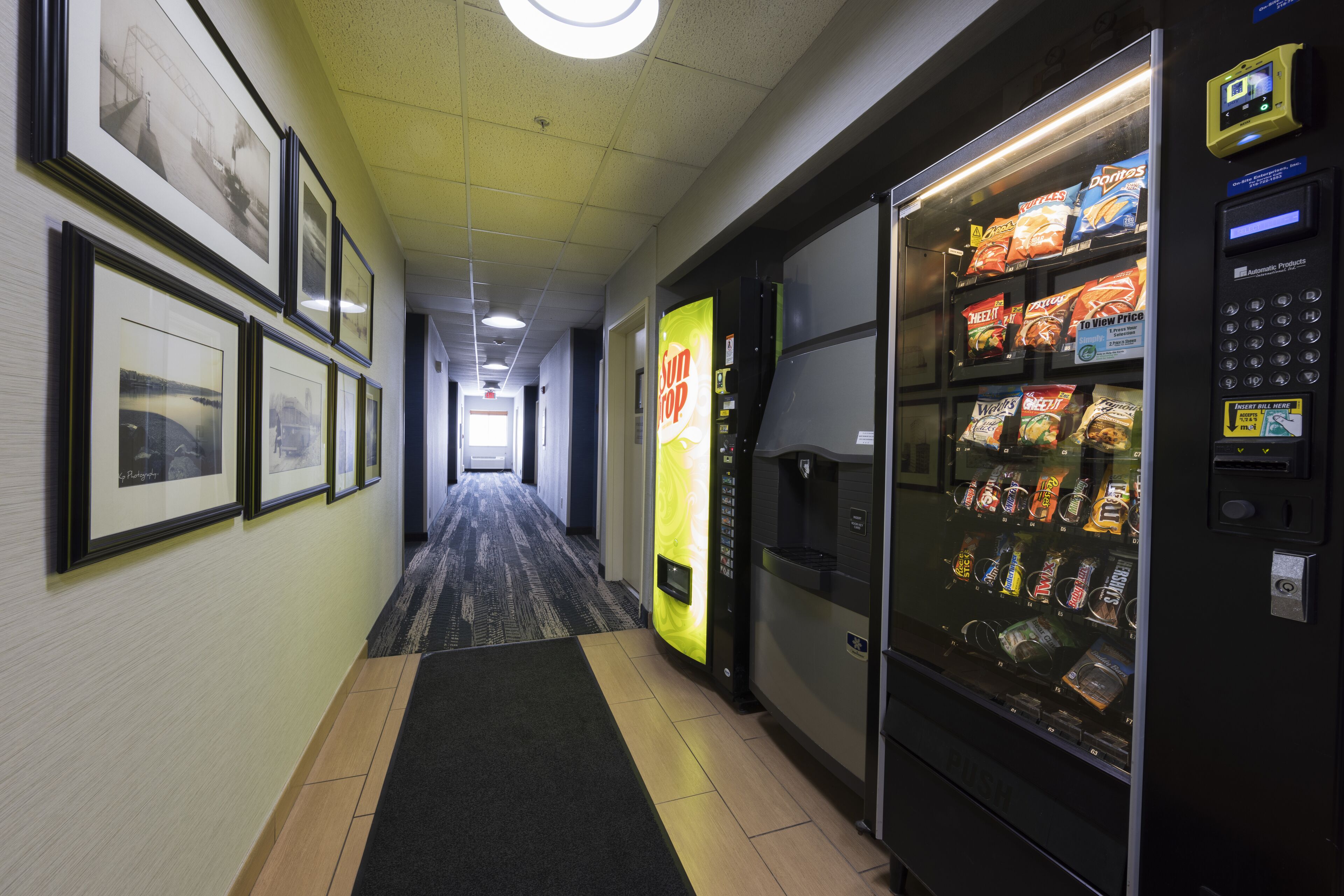 A hotel hallway with framed pictures on the wall, a vending machine with snacks, and a carpeted floor.