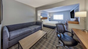 A hotel room with a gray sofa, a single bed, a desk with a chair, a lamp, and a TV.