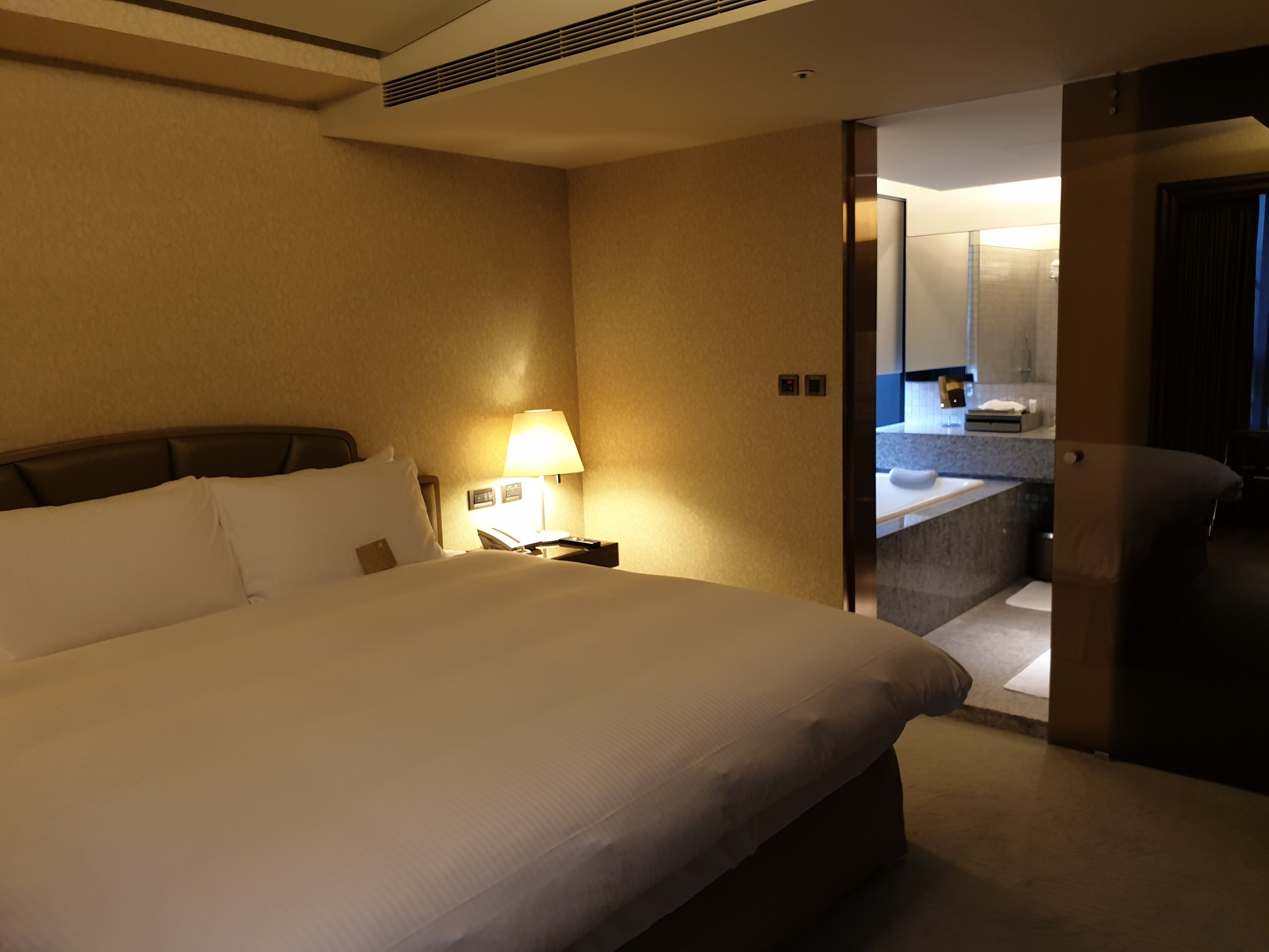 A hotel room with a large bed, a bedside lamp, a nightstand, and a view of the bathroom through an open door.