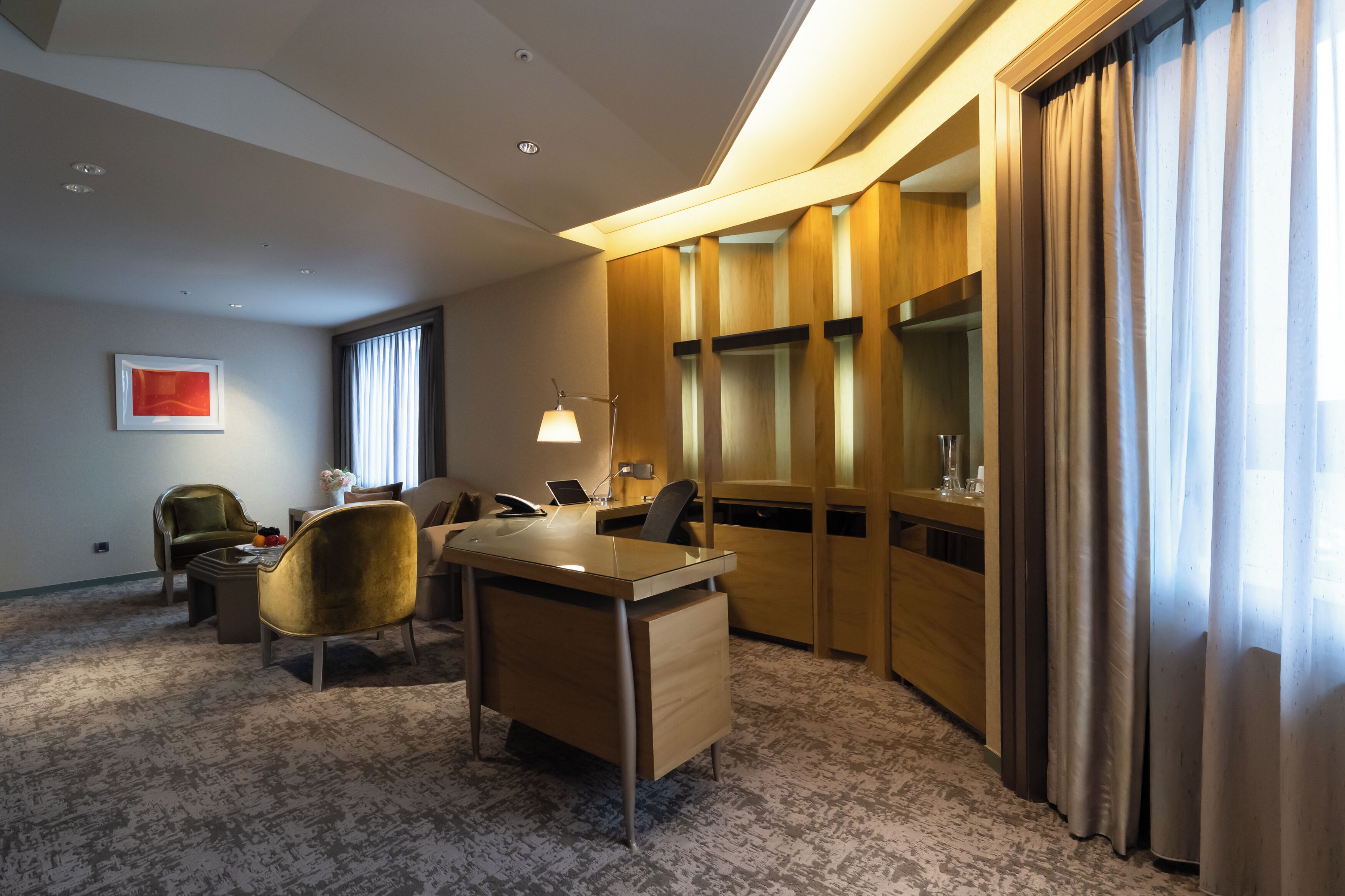 Una habitación de hotel con escritorio, dos sillas, una lámpara, una mesita con un jarrón y un ventanal con cortinas.