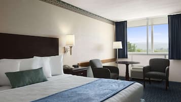 A hotel room with a large bed, a desk with a lamp, two chairs, a small table, and a view of the outdoors through a window.