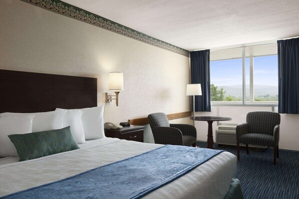 A hotel room with a large bed, a desk with a lamp, two chairs, a small table, and a view of the outdoors through a window.