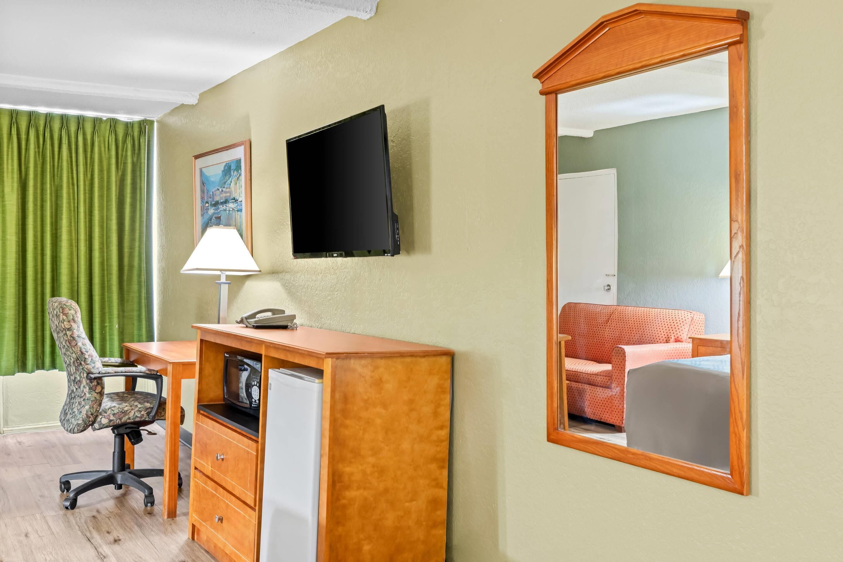 A hotel room with a wooden desk, a chair, a television, a refrigerator, a mirror, and a green curtain.
