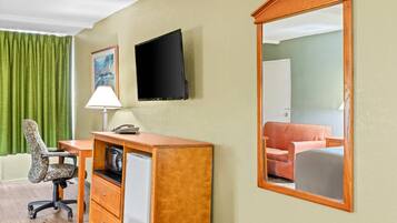 A hotel room with a wooden desk, a chair, a television, a refrigerator, a mirror, and a green curtain.