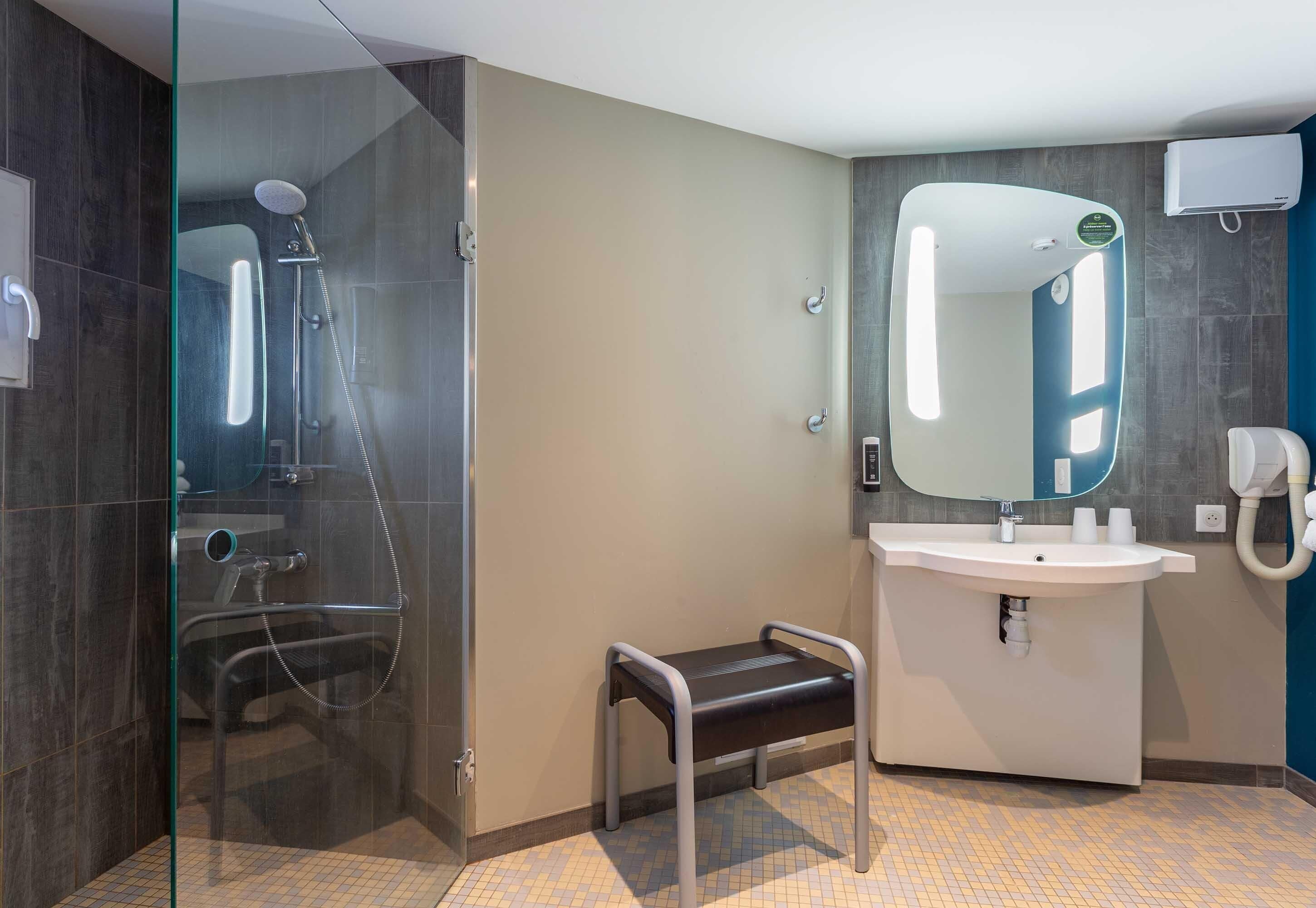 Un baño moderno con ducha envuelta en cristal, un lavamanos blanco, un espejo y un pequeño banco.