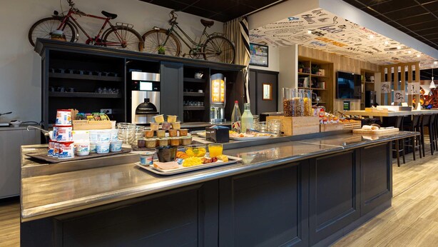 A breakfast bar with various food items, a bicycle mounted on the wall, and a wall with drawings. - B&B Hotel Orléans Centre Foch (Orléans)