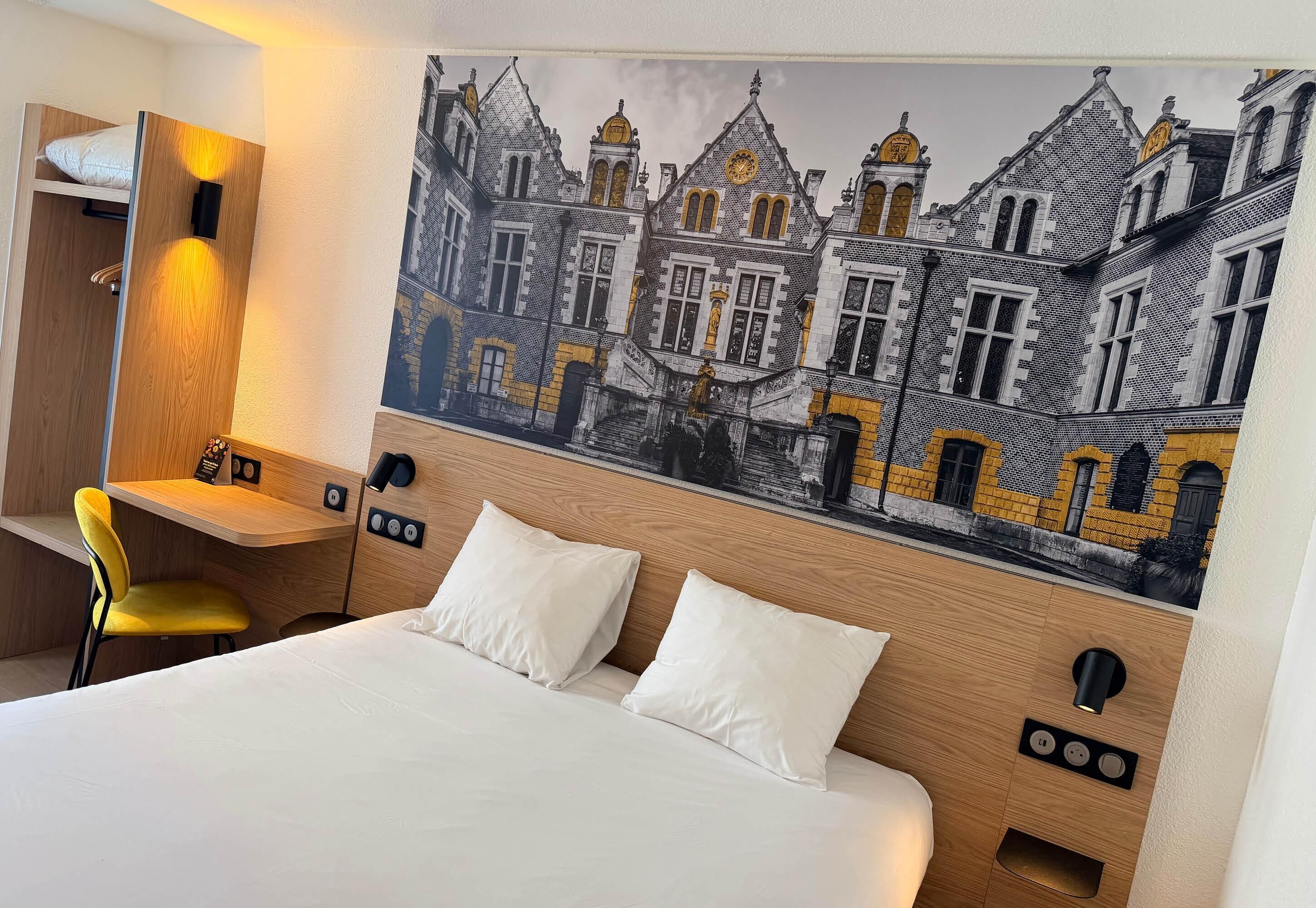 A hotel room with a bed, a desk, a chair, and a large wall mural of buildings.