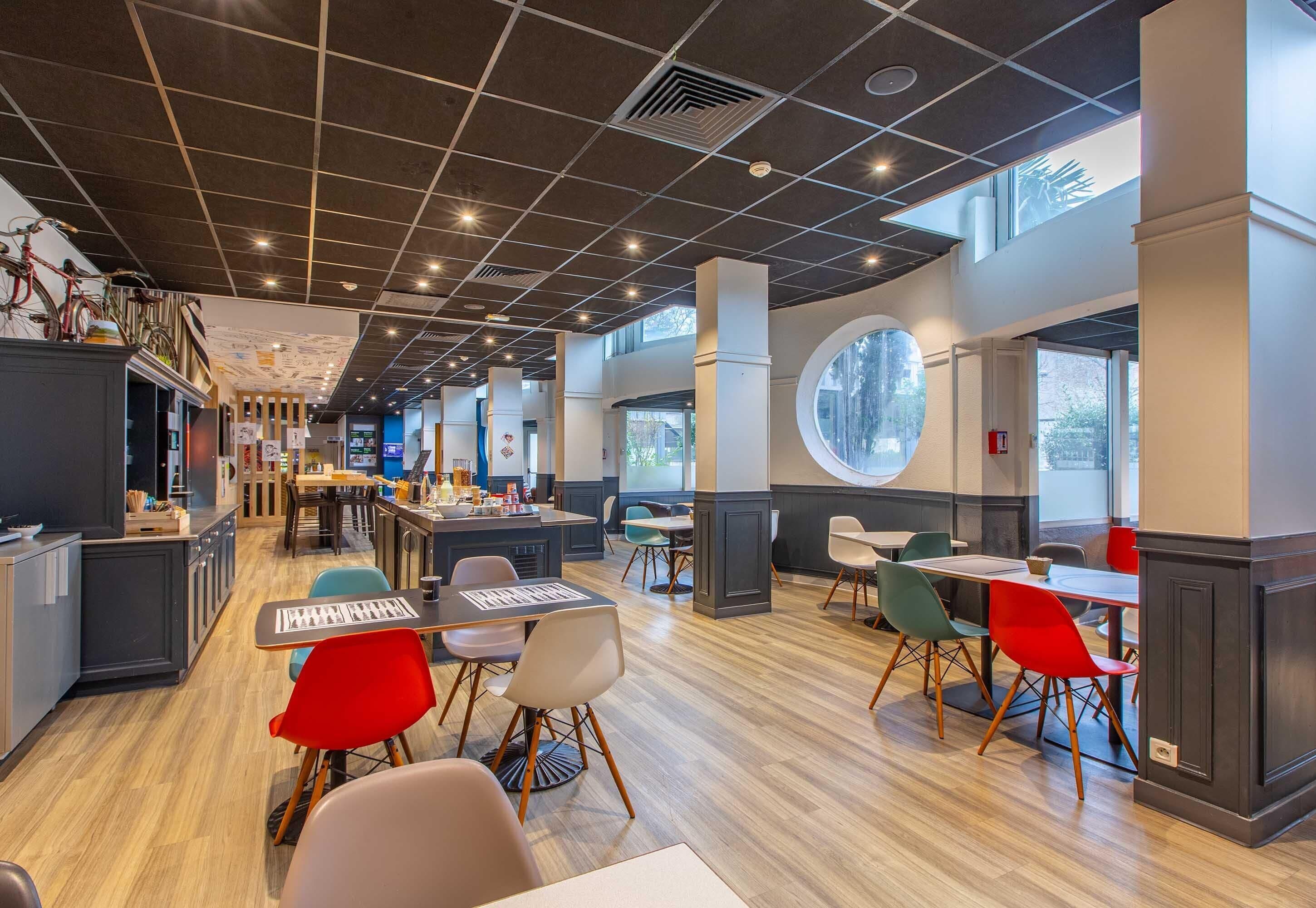 A modern restaurant with a high ceiling, wooden flooring, and a mix of red and white chairs.