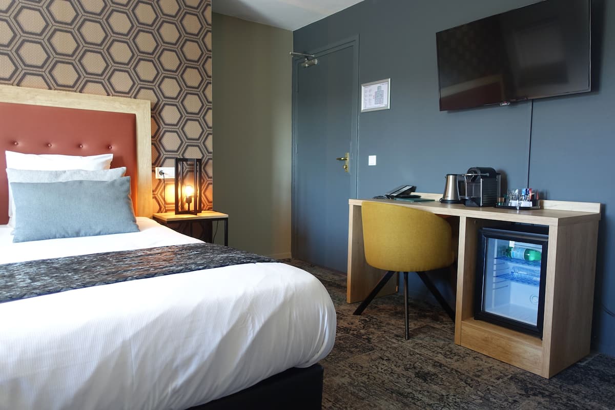Habitación de hotel con una cama, un escritorio con computadora, una cafetera y un televisor en la pared.