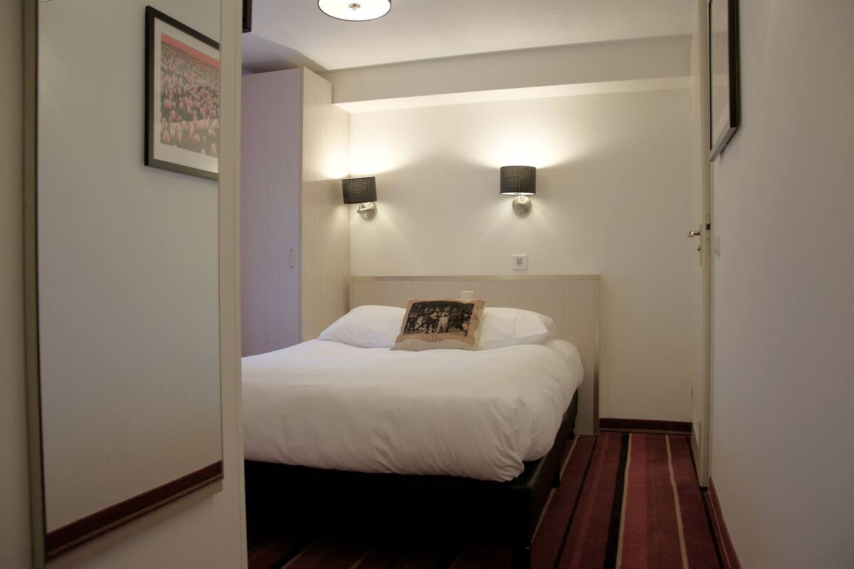 Habitación de hotel con cama, dos lámparas de pared, un cuadro enmarcado y suelo cubierto de moqueta roja.