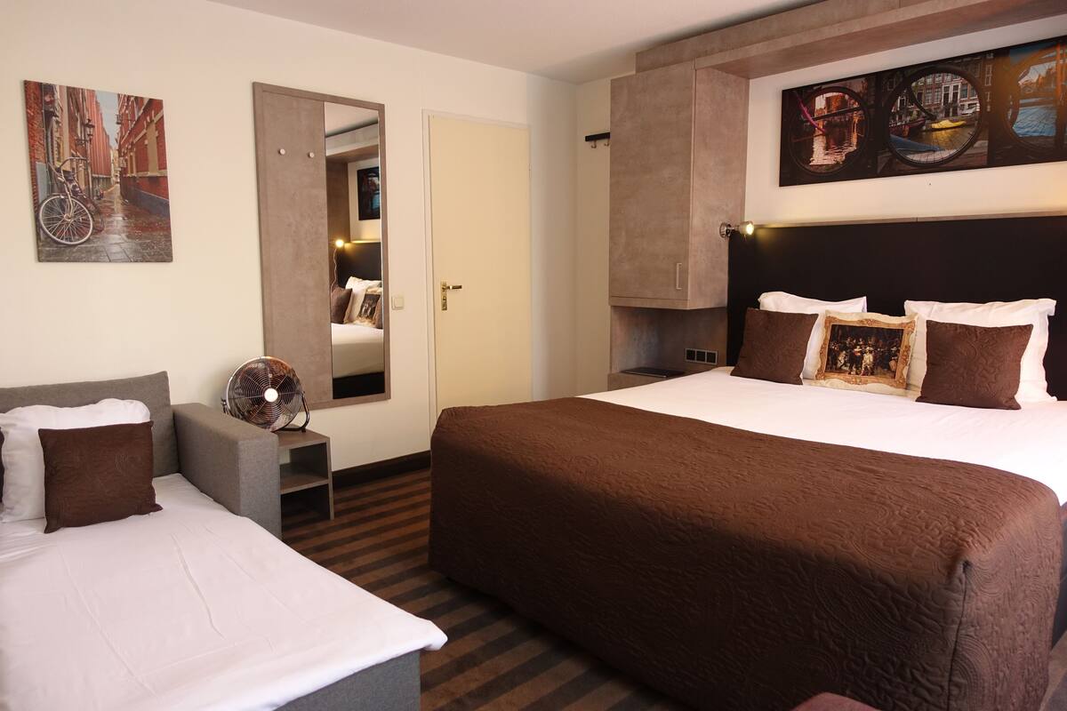 Una habitación de hotel con una cama grande, una cama más pequeña, una zona de estar, un espejo y obras de arte en las paredes.