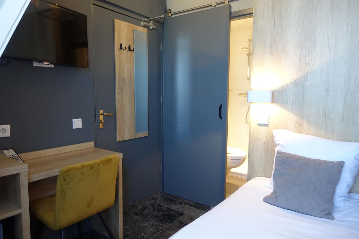 Habitación de hotel con cama, escritorio con silla, espejo, puerta y baño visible a través de una puerta abierta.