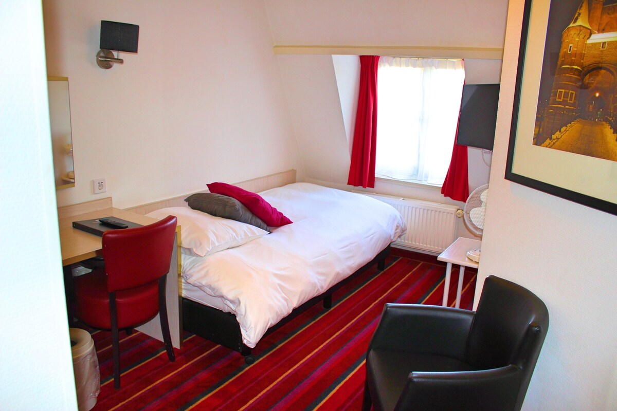 Camera d'albergo con un letto, una scrivania, una sedia, un termosifone e una finestra con tende.