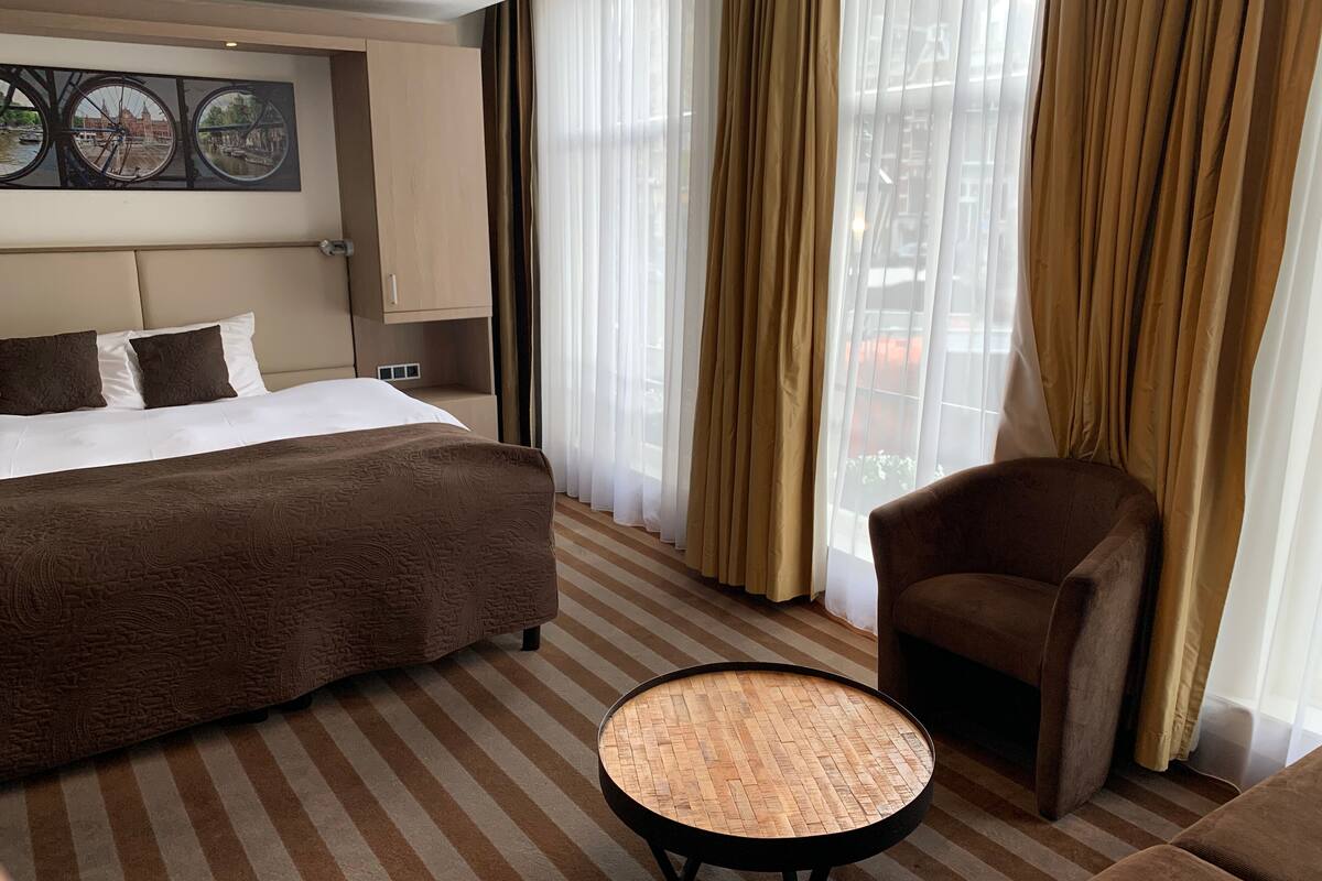 Habitación de hotel con una cama grande, una mesa redonda de madera, un sillón marrón y vistas a un puente por la ventana.