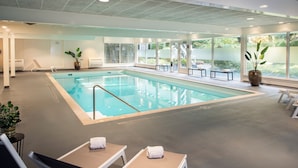 A large indoor swimming pool with clear glass walls, lounge chairs, and potted plants. - Bilderberg Hotel 't Speulderbos (Garderen)