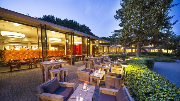 An outdoor dining area with wooden tables and chairs, surrounded by a hedge and a building with large windows. - Bilderberg Hotel 't Speulderbos (Garderen)