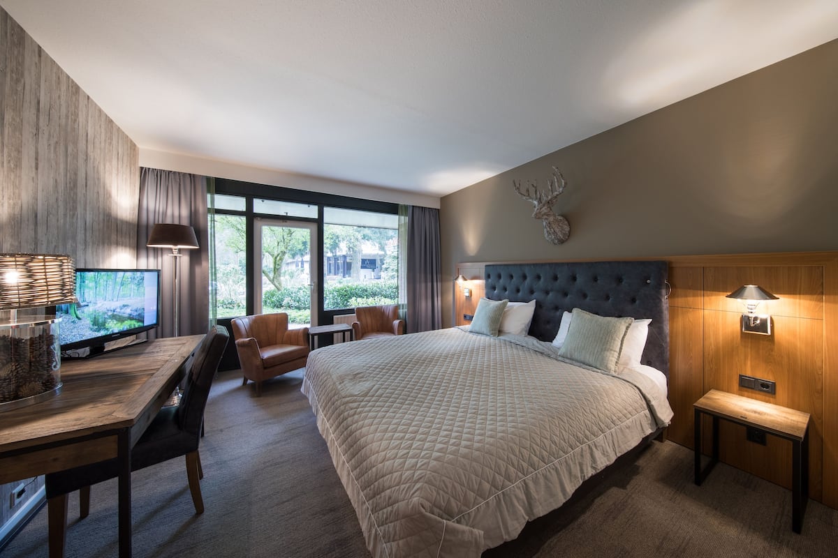 A modern hotel room with a large bed, a desk with a TV, and a view of the outdoors.