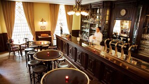 Zona de bar con mesas y sillas de madera, un barman y una barra bien surtida.