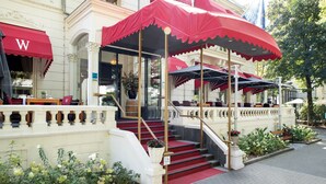 Entrada de hotel con toldos rojos, una escalera y mobiliario exterior.