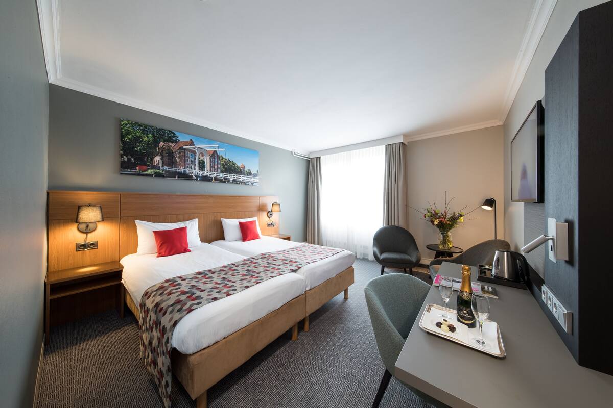 A hotel room with a large bed, bedside tables with lamps, a desk with a tray of champagne, and a chair.