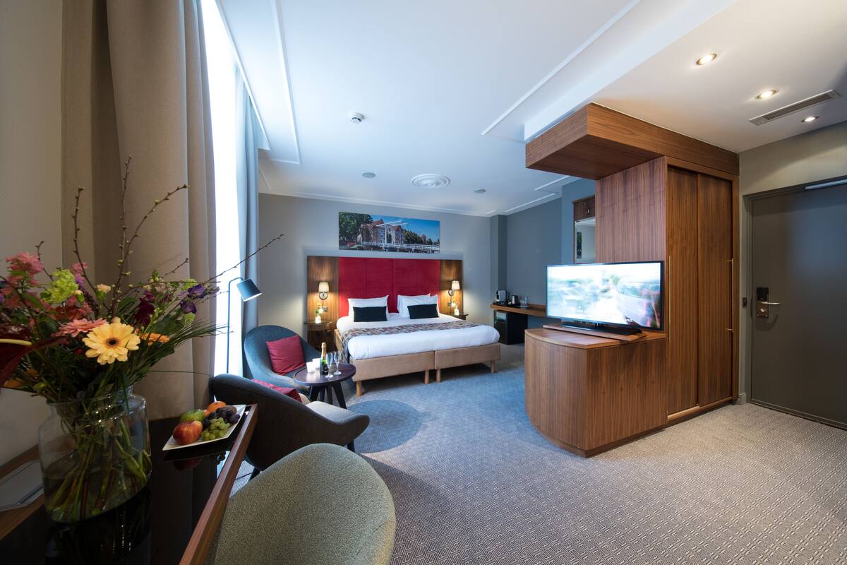 A hotel room with a bed, a TV, a desk with a vase of flowers, and a chair.