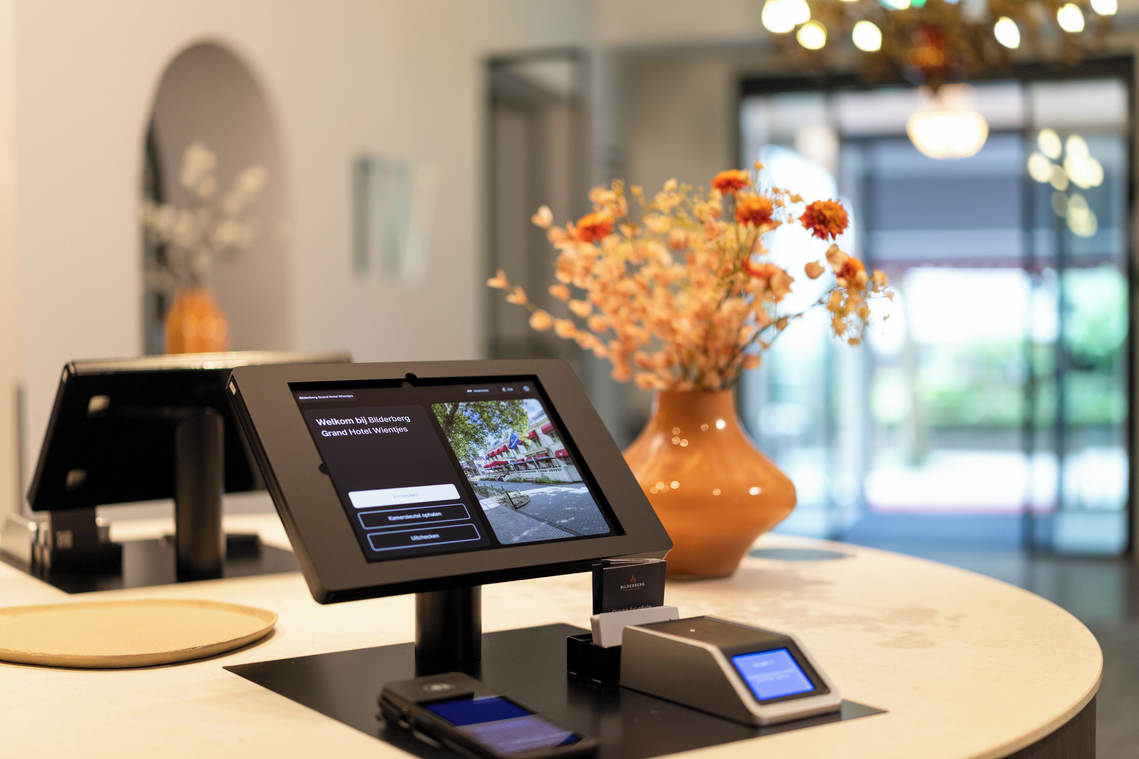 A hotel reception area with a digital kiosk, a vase of flowers, and a tablet displaying a welcome message.