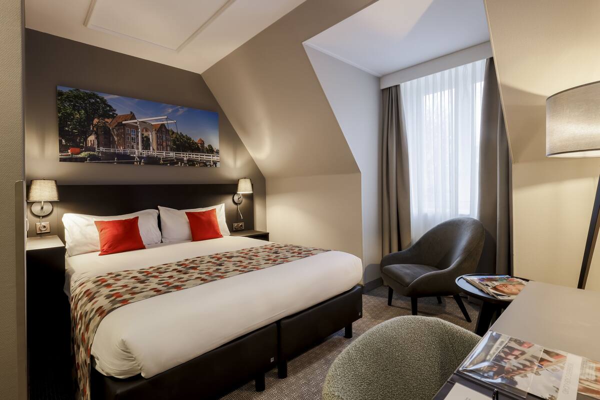 A hotel room with a large bed, a chair, a desk, and a window with curtains.