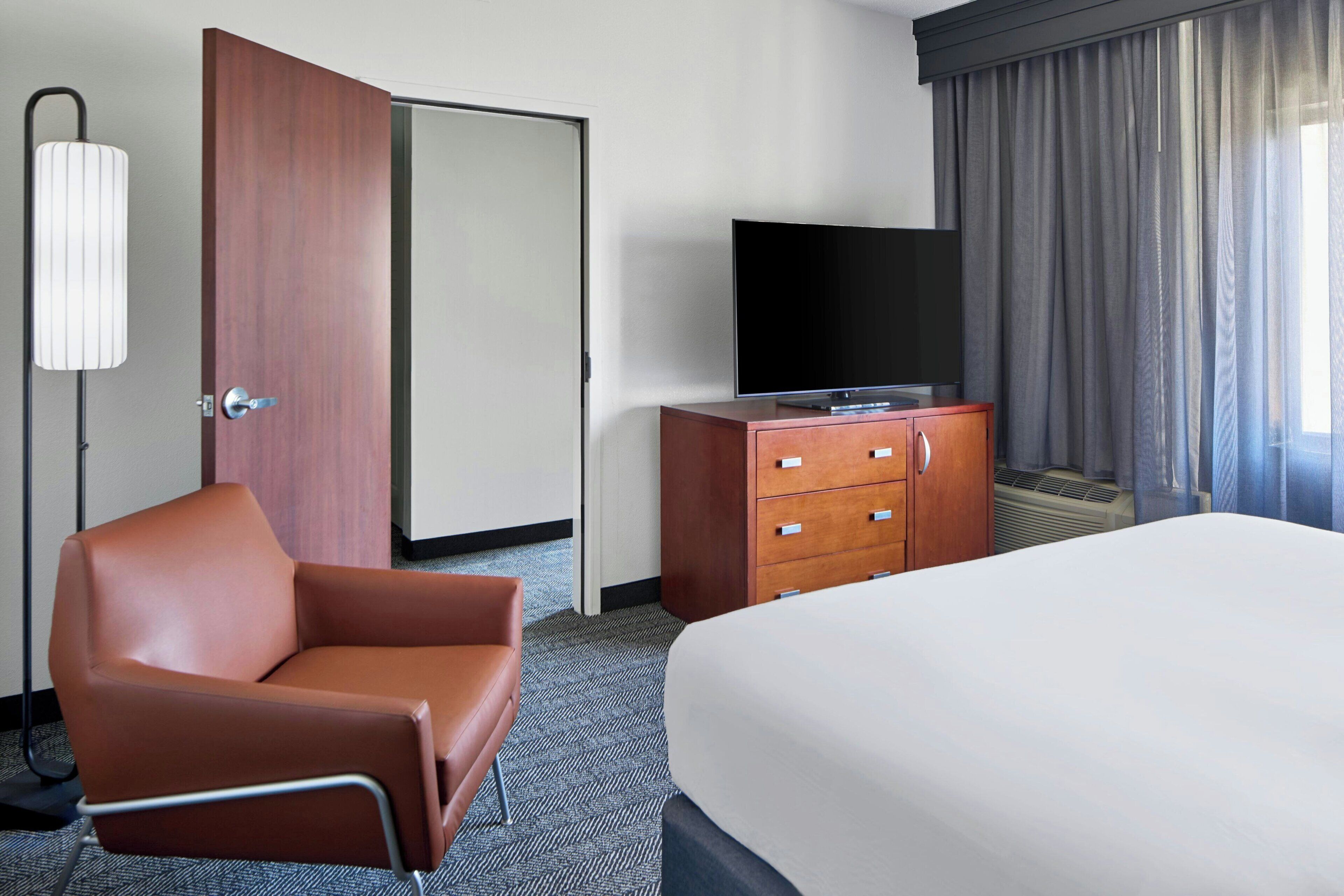 A hotel room with a bed, a television on a wooden cabinet, a brown leather chair, and a closed door.