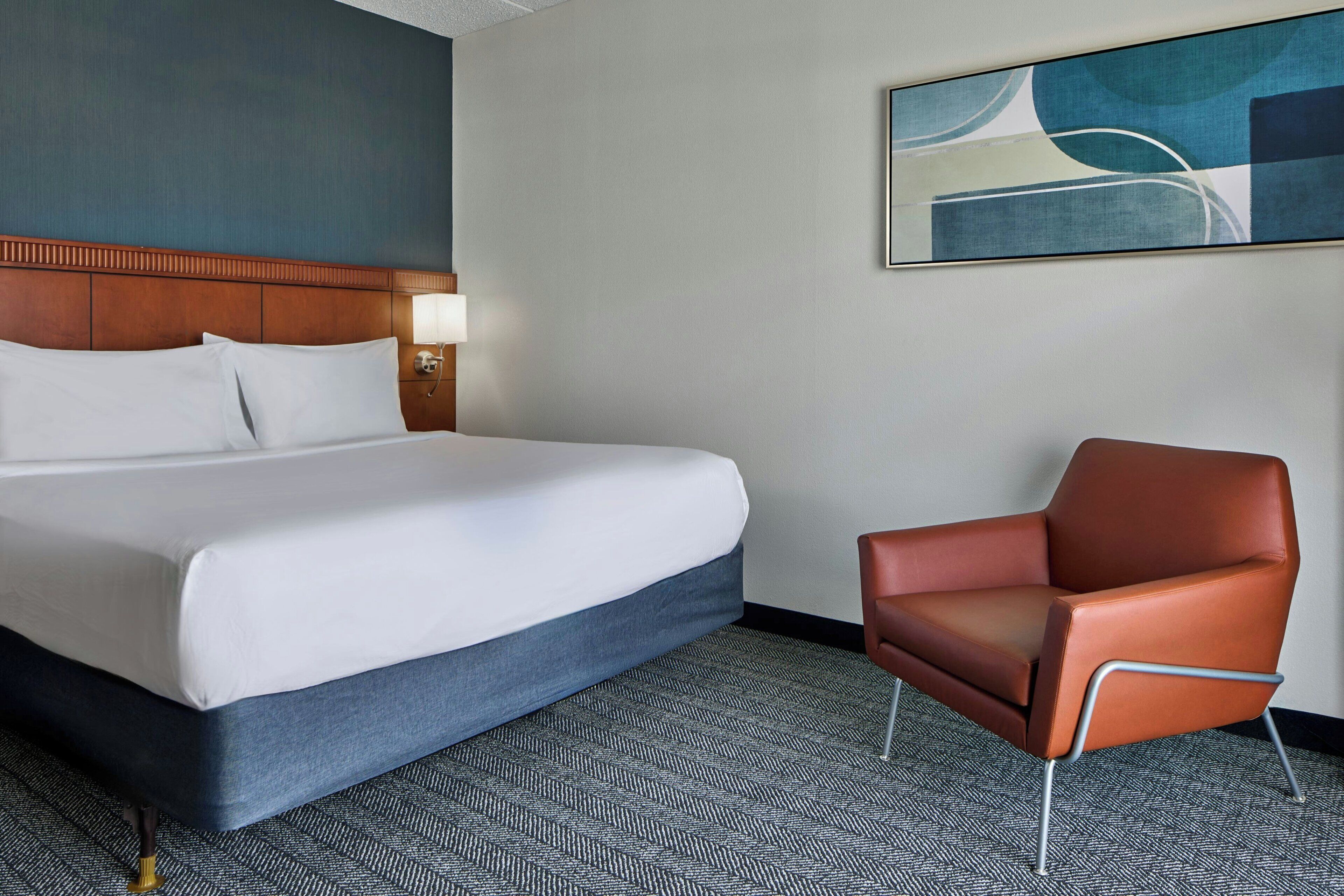 A hotel room with a large bed, a chair, a headboard, a wall-mounted lamp, and a painting.