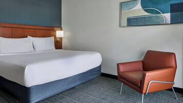 A hotel room with a large bed, a chair, a headboard, a wall-mounted lamp, and a painting.