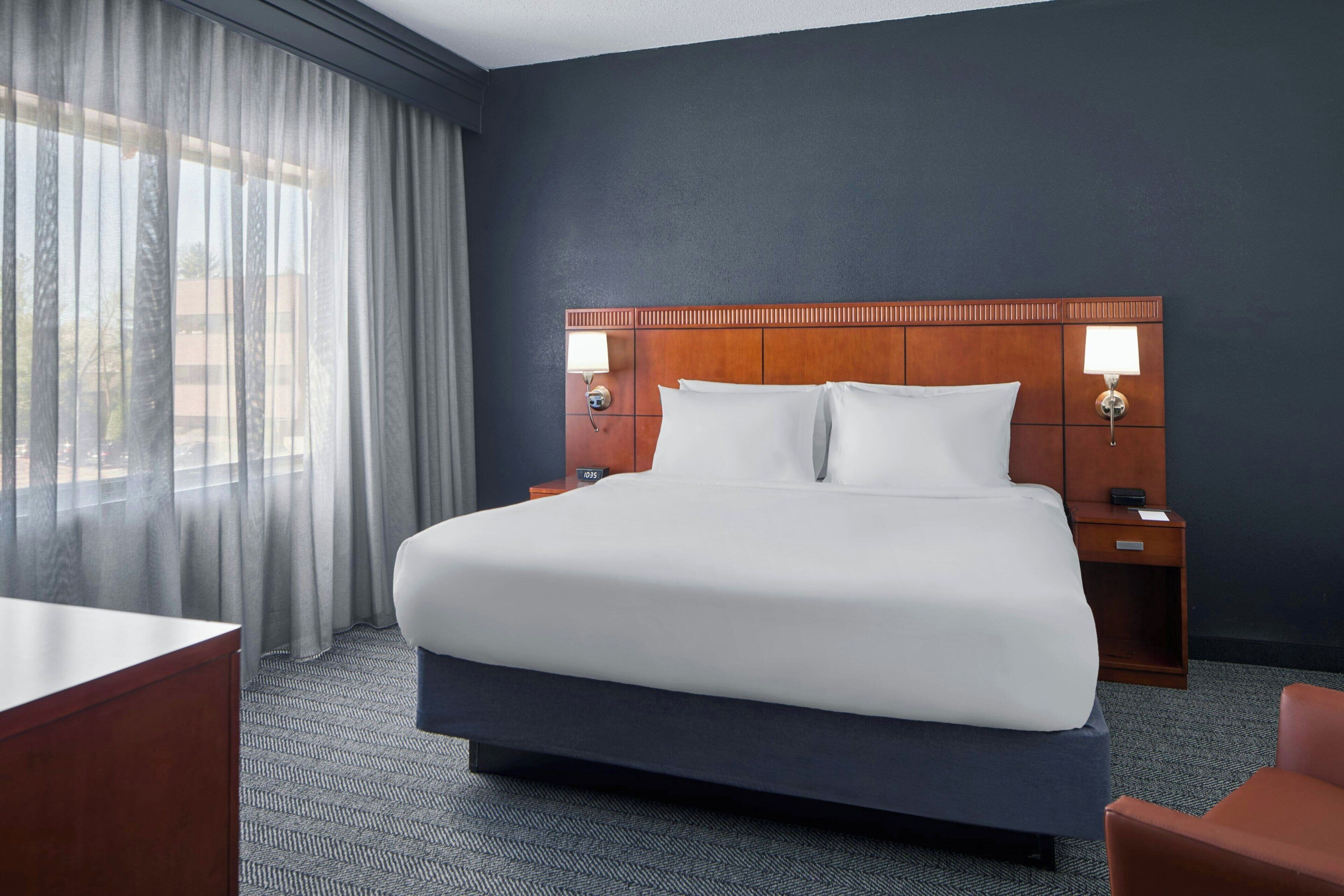 A hotel room with a large bed, two bedside tables with lamps, a desk, a chair, and a view through sheer curtains.