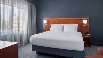 A hotel room with a large bed, two bedside tables with lamps, a desk, a chair, and a view through sheer curtains.