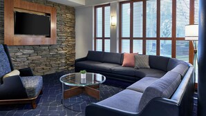 A modern living room with a stone wall, a large flat-screen TV, a sectional sofa, a glass coffee table, and large windows with blinds. - Courtyard by Marriott Rochester Brighton (Rochester)