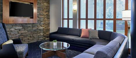 A modern living room with a stone wall, a large flat-screen TV, a sectional sofa, a glass coffee table, and large windows with blinds.