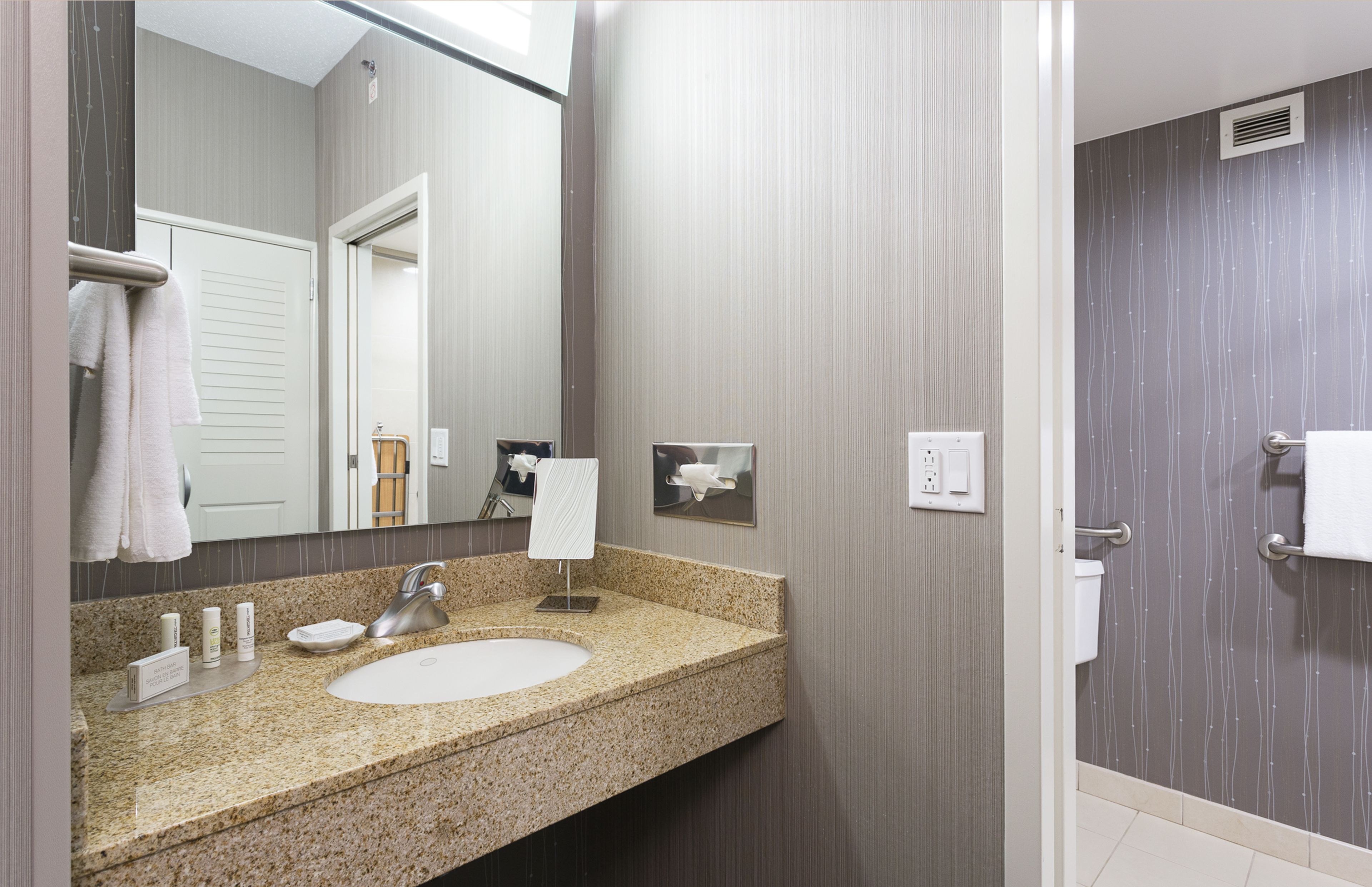 A modern bathroom with a granite countertop sink, a large mirror, and a shower area with a towel rack.