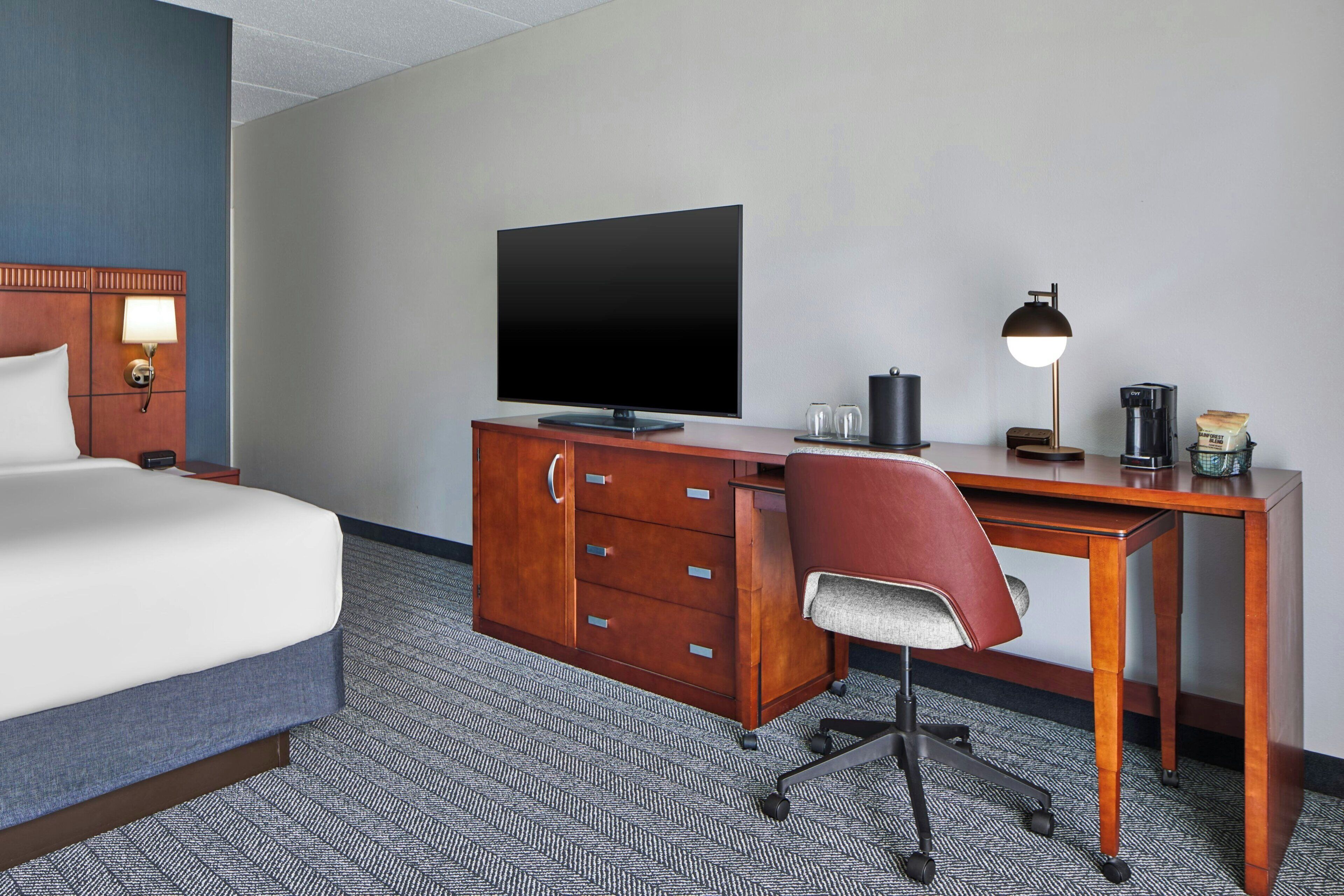 Una habitación de hotel con cama, escritorio, silla, televisor, lámpara y cafetera.