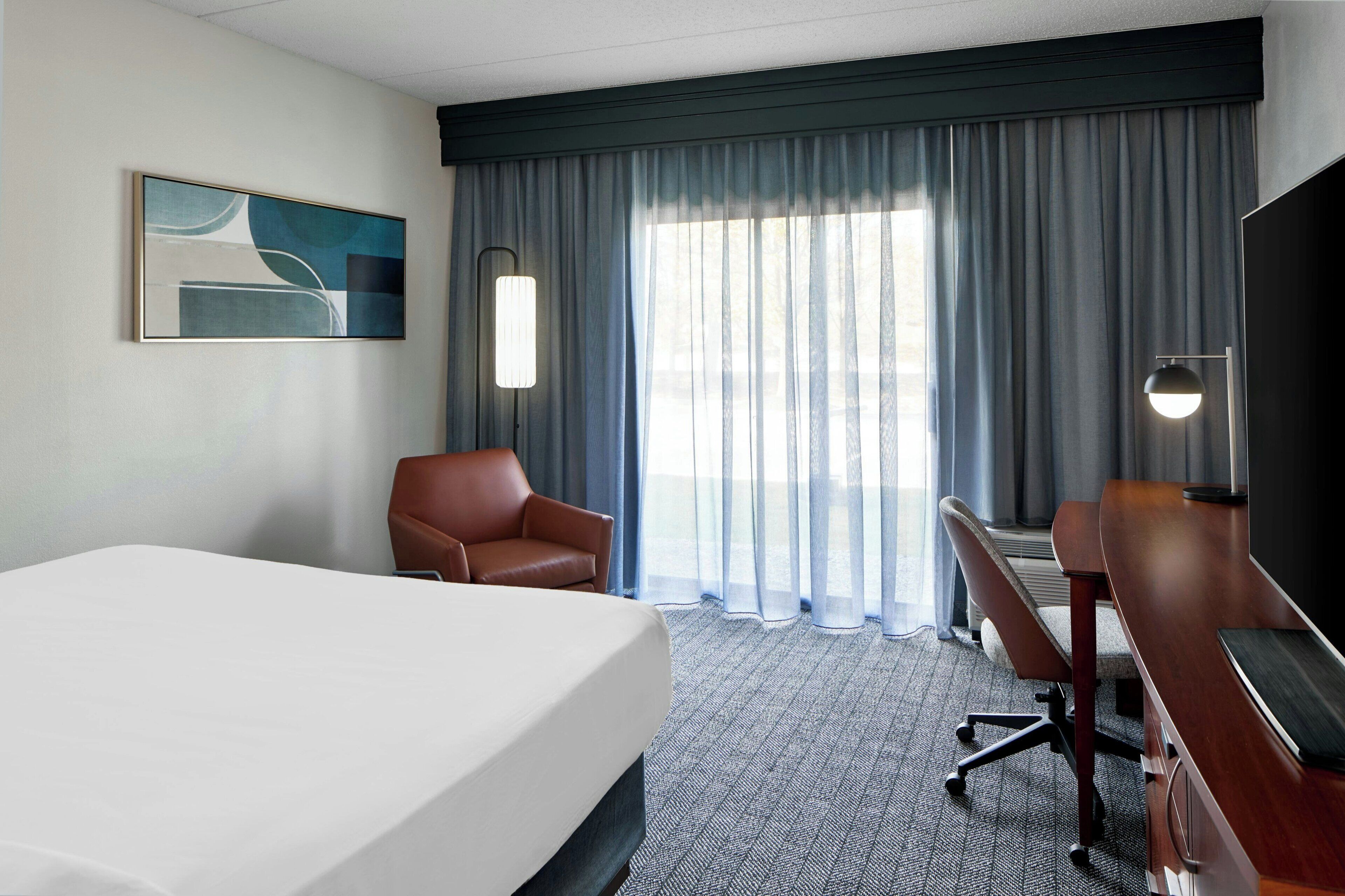 A hotel room with a bed, a desk, a chair, a TV, and a large window with curtains.