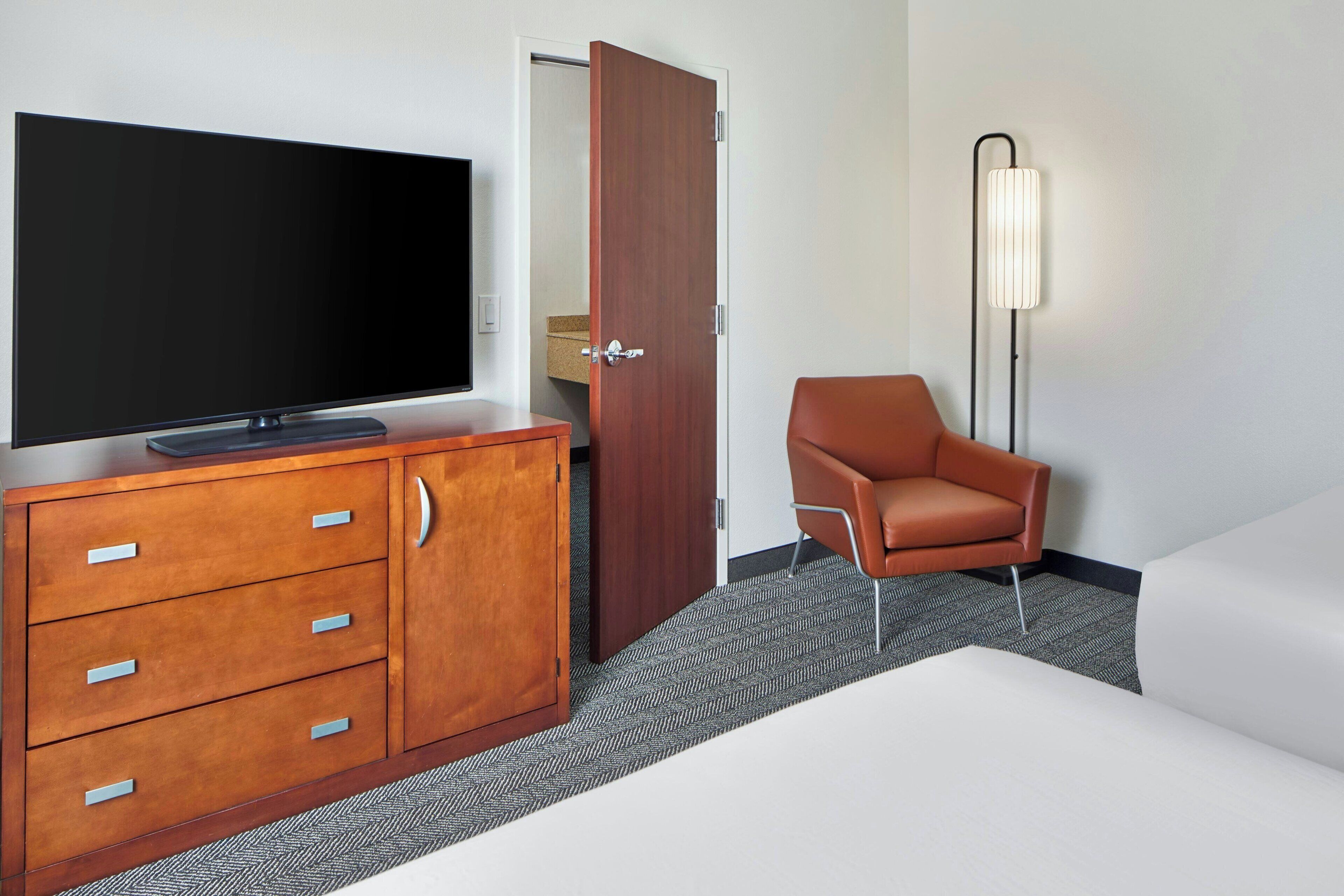 A hotel room with a flat-screen TV, a wooden dresser with drawers, a brown chair, a white sofa, a floor lamp, and a door.