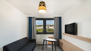 A modern hotel room with a sofa, a small table, a TV mounted on the wall, and a window offering a city view. - Mercure Budapest Castle Hill (Budapest)