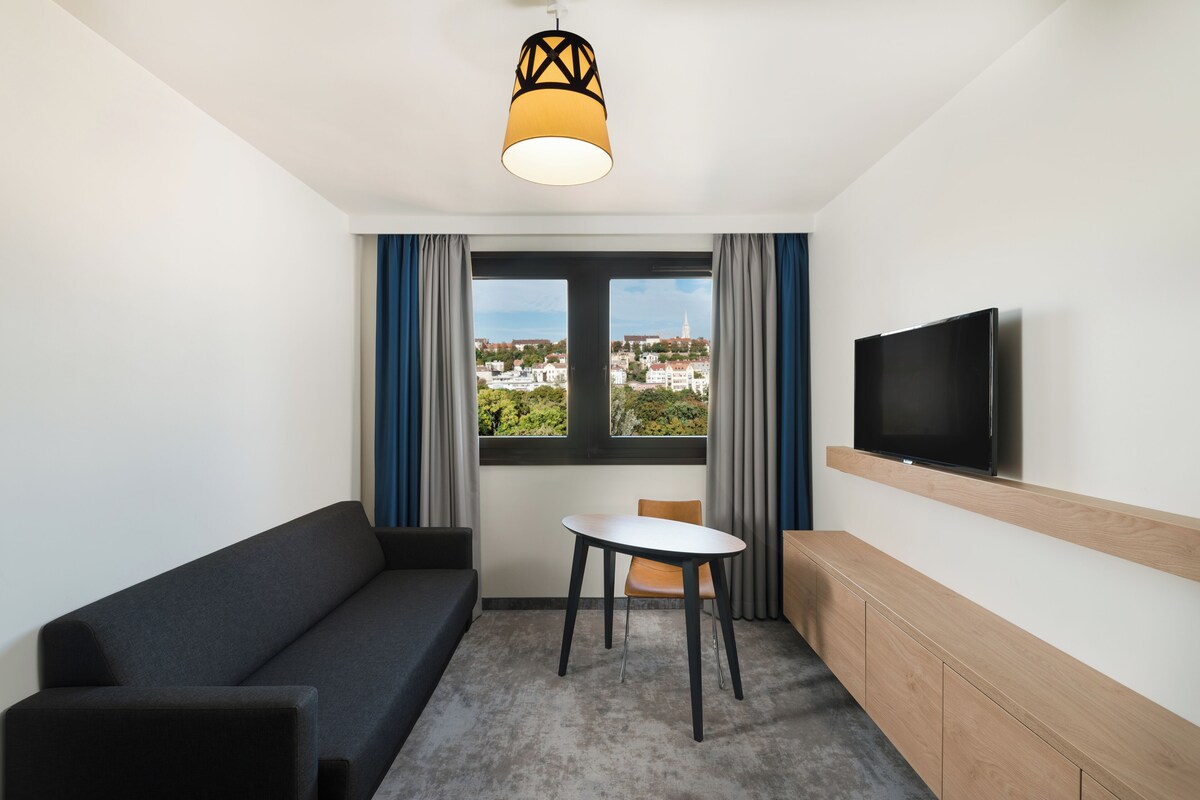 Una habitación de hotel moderna con sofá, una mesita, televisor montado en la pared y ventana con vistas a la ciudad.
