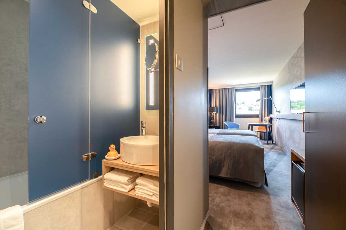 Habitación de hotel con baño que incluye una bañera, un lavabo y un espejo. La habitación tiene una cama, un escritorio y una ventana con cortinas.