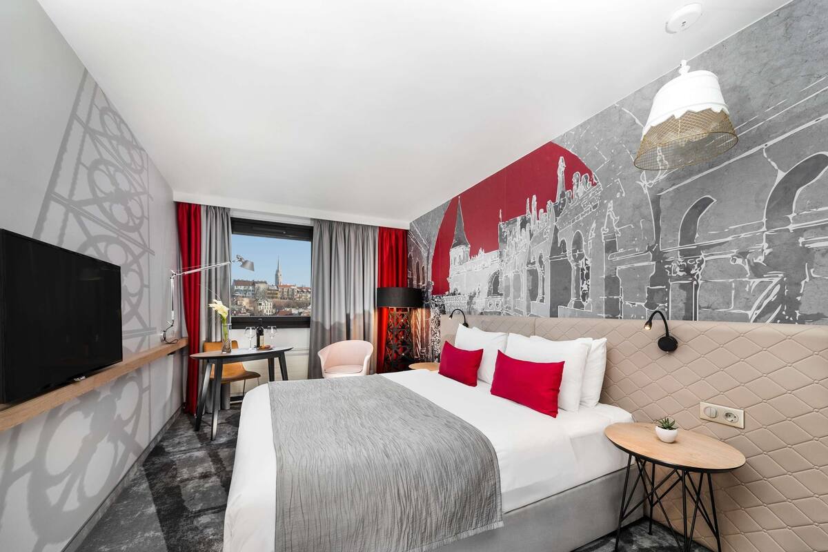 A hotel room with a bed, a TV, a desk with a chair, a small table, and a wall mural.