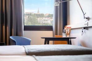 A hotel room with a bed, a desk with a lamp, a chair, and a view of a cityscape through a window. - Mercure Budapest Castle Hill (Budapest)