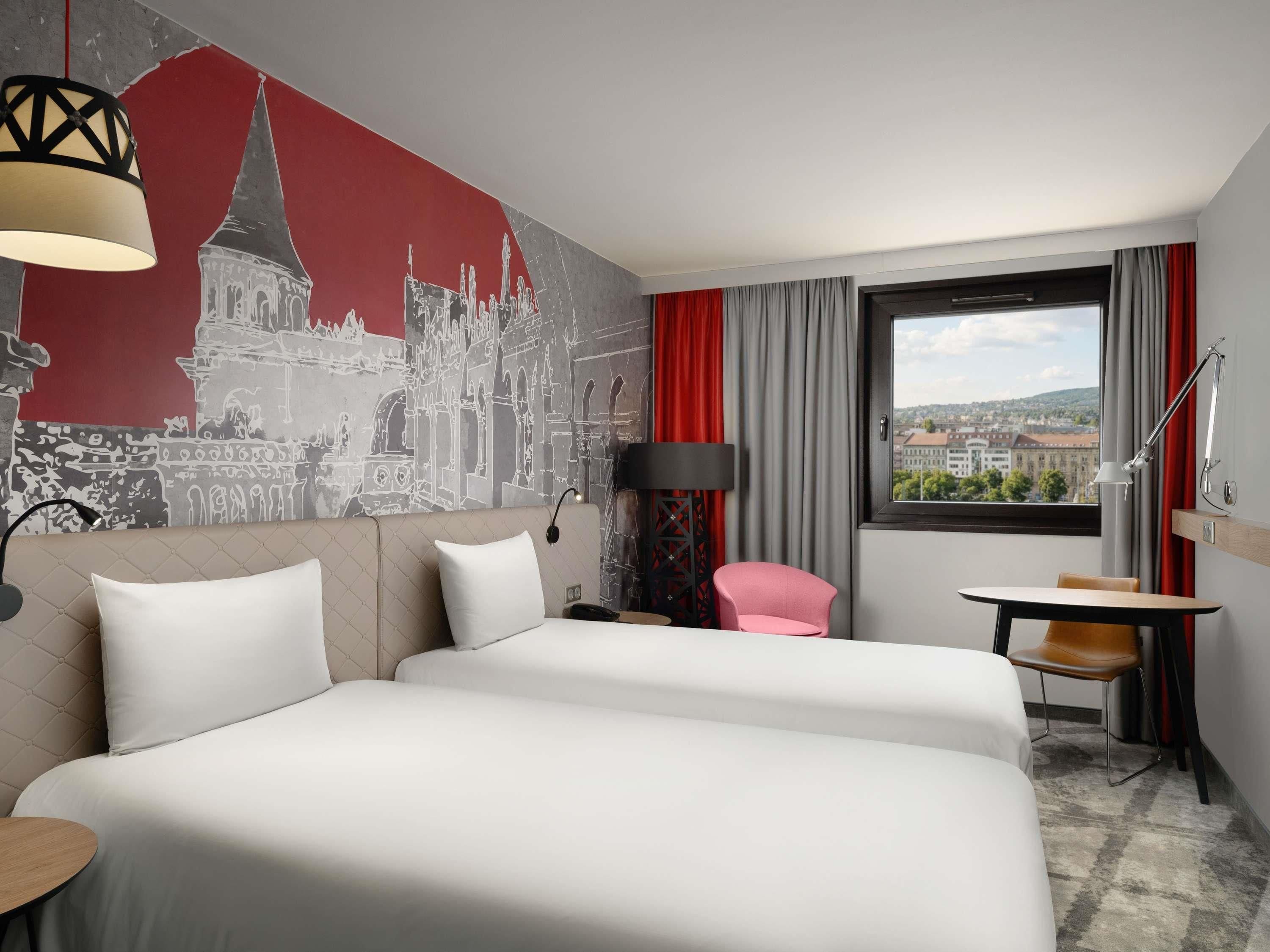 Habitación de hotel con dos camas, un mural con vista a la ciudad, un escritorio con silla, una ventana y una cortina roja.