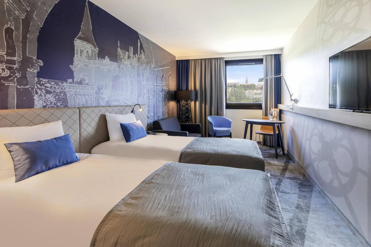 A hotel room with two beds, a desk, a chair, a lamp, and a view of a cityscape.