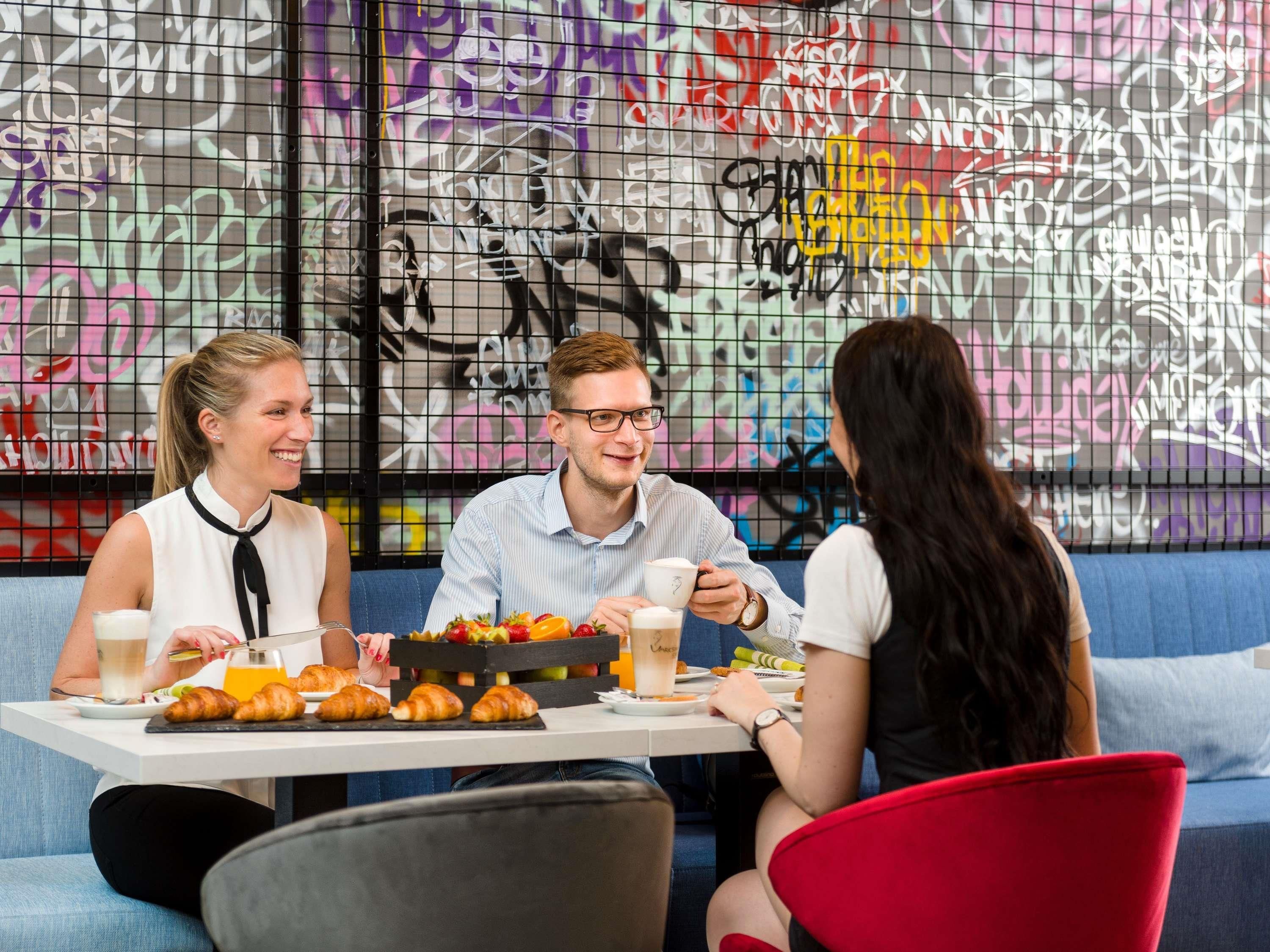 Um grupo de pessoas sentadas à mesa, com comida e bebidas, em um ambiente com grafite na parede.