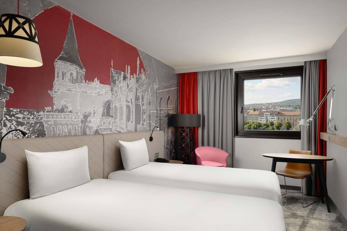 A hotel room with two beds, a cityscape mural, a desk with a chair, a window with a view, and a red curtain.